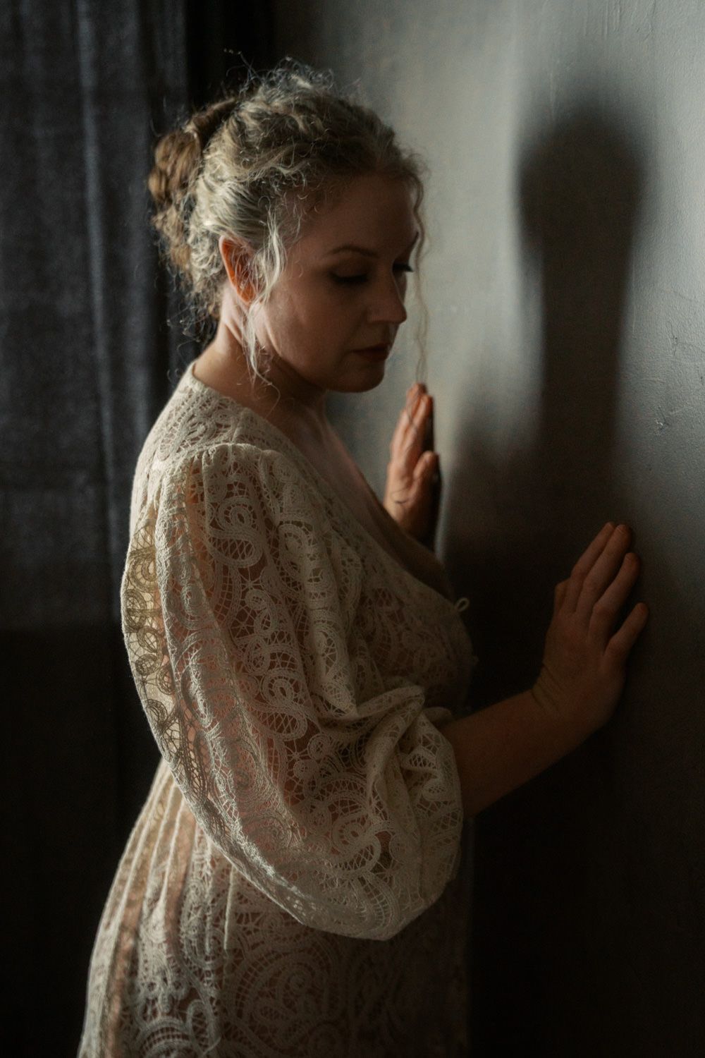 Woman in lace robe leaning against dark wall with dramatic shadow, moody portrait photography.
