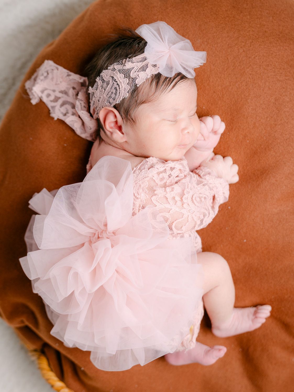 Sleeping newborn baby girl in pink lace ruffle dress and floral headband, resting on a brown cushion.