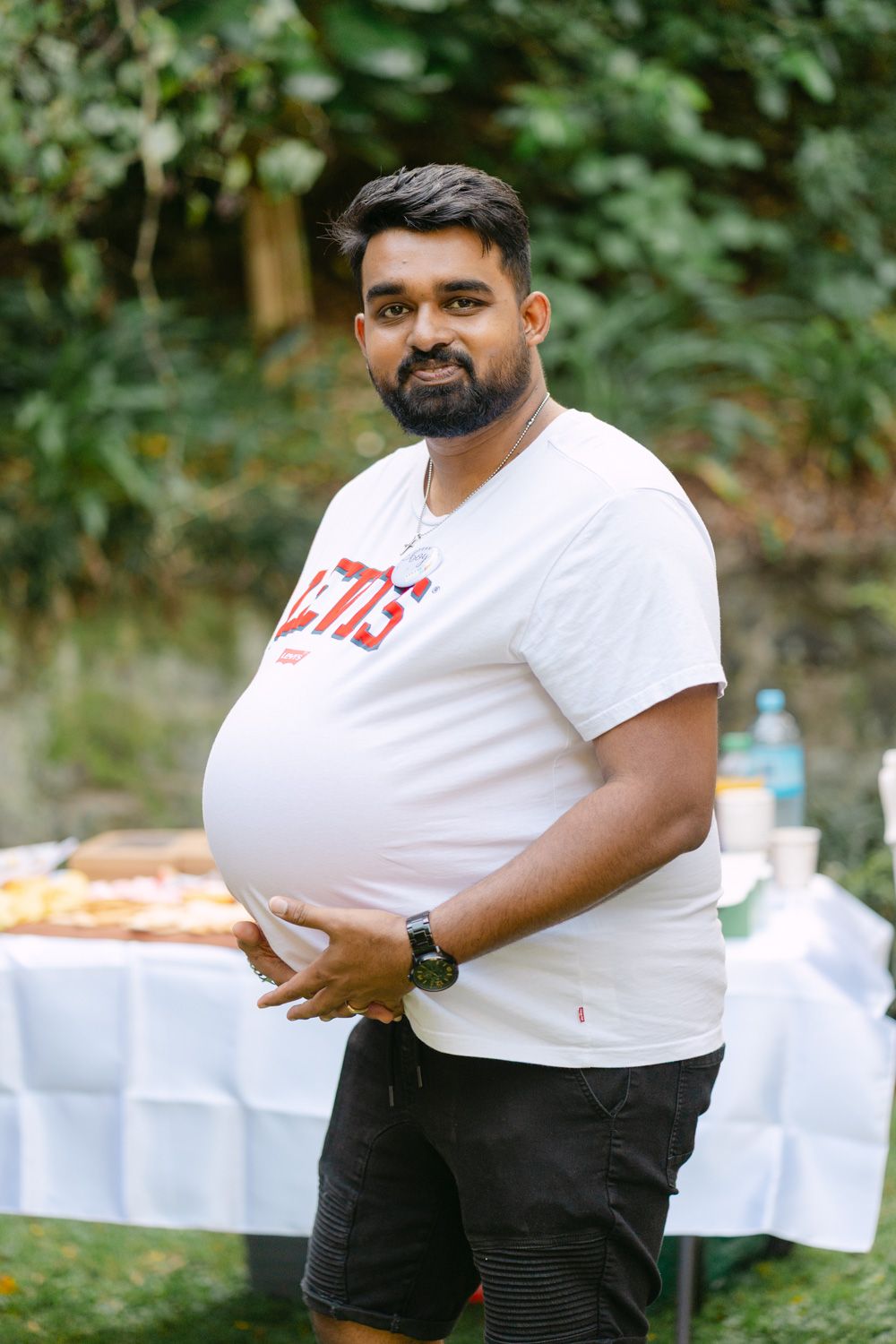 A bearded man in a white graphic t-shirt stands outdoors near a food table, posing for a photo.