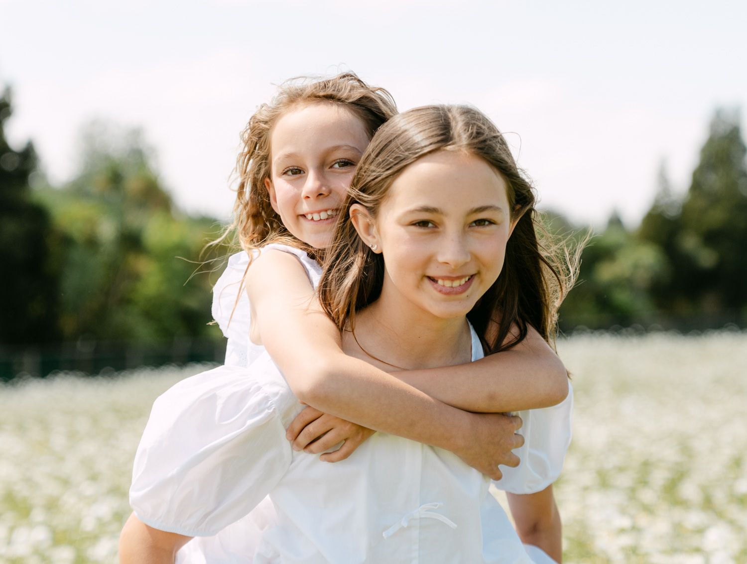 Two smiling girls in white dresses enjoy a piggyback ride in a sunny flower field.