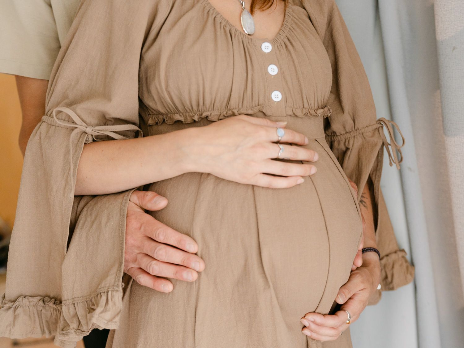 Pregnant woman in beige dress cradling baby bump with partner's hands gently resting on her belly.