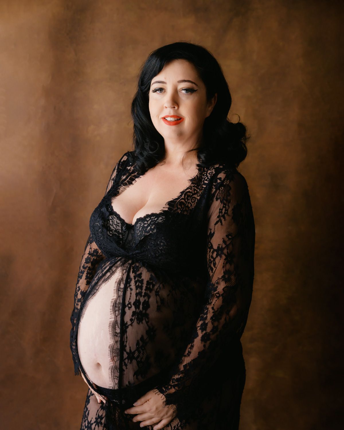 Pregnant woman in black lace maternity gown posing elegantly against a brown studio backdrop.