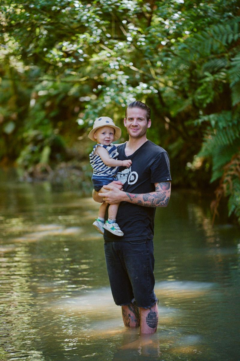 Tattooed man holding a toddler in a bucket hat while standing in a shallow creek surrounded by lush green trees.