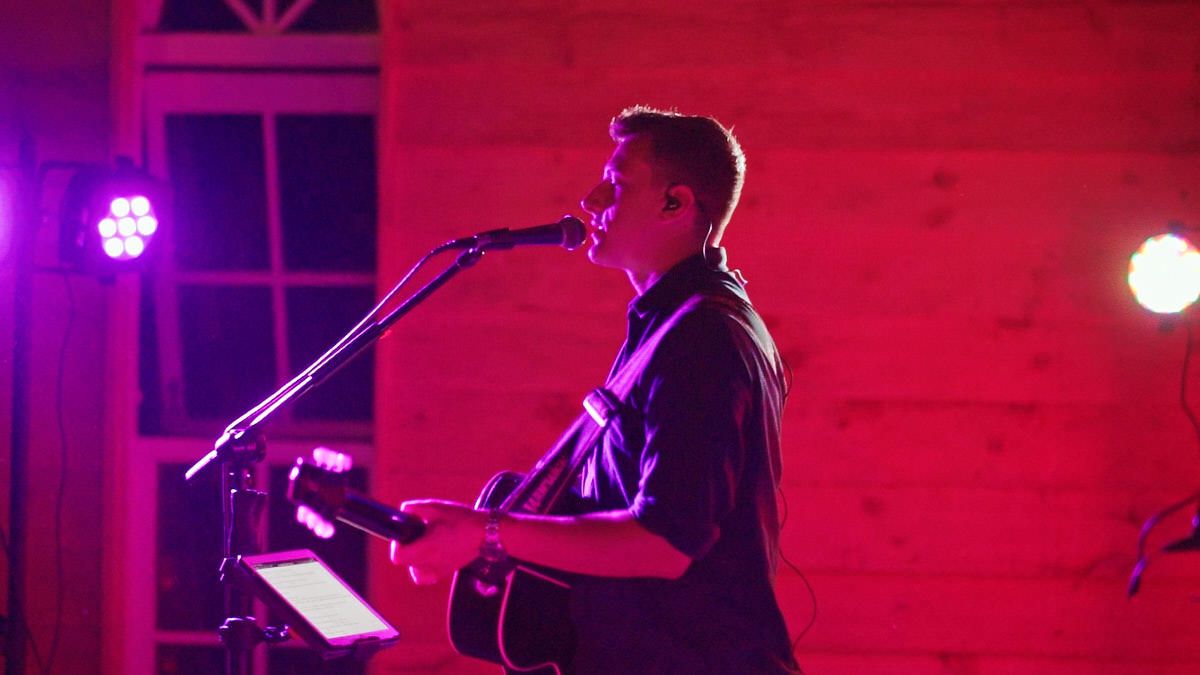 Male guitarist performing on stage under pink and purple stage lights at an indoor venue.
