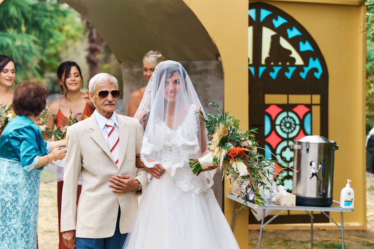 Bride in white gown and veil walks down the aisle with an elderly man in a suit at an outdoor wedding ceremony.