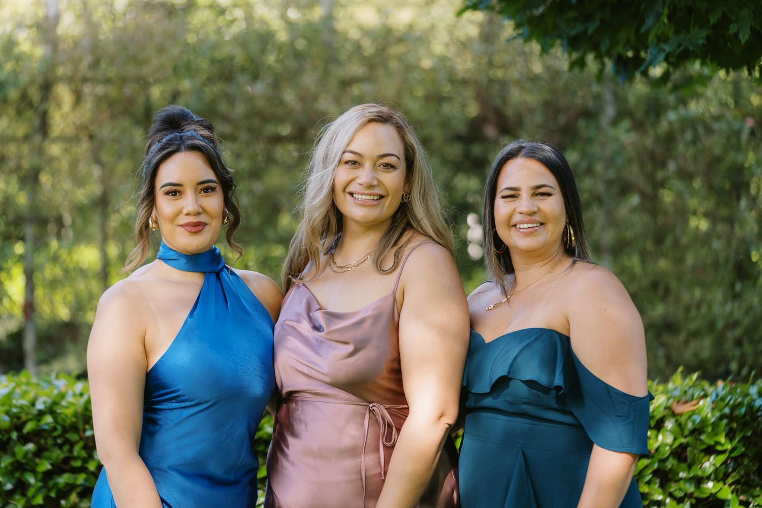 Three women in elegant dresses smiling together outdoors, surrounded by lush green foliage.