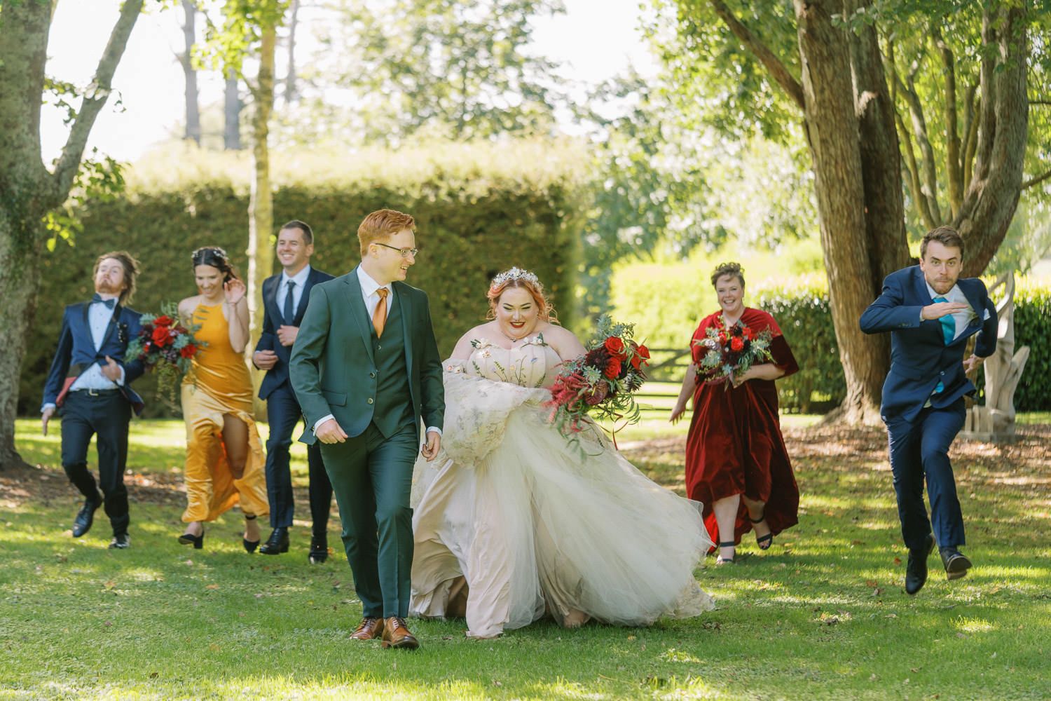 Bride and groom walk joyfully with wedding party on green lawn, bridesmaids in jewel tones holding red bouquets.