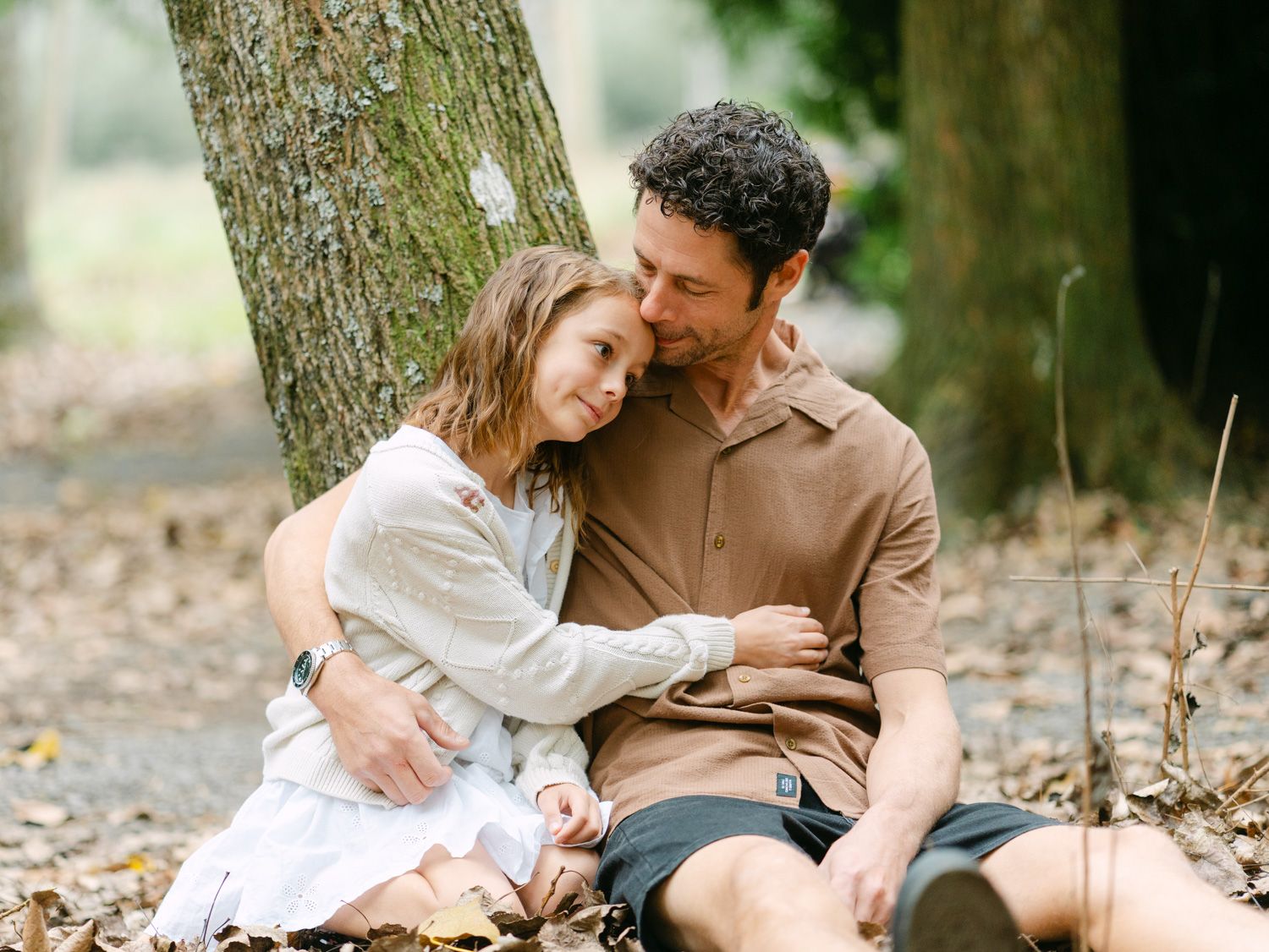 Couple sitting together against a tree trunk in autumn leaves, sharing a tender romantic embrace outdoors.