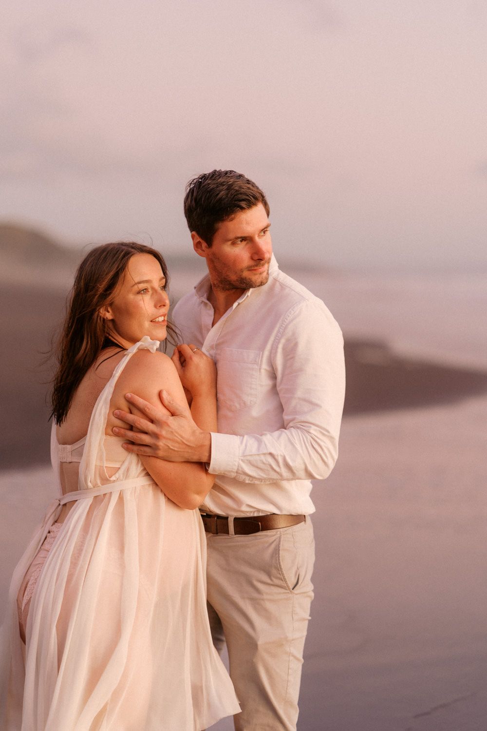 Romantic couple embracing on a beach at sunset, woman in white dress, man in white shirt and khaki pants.
