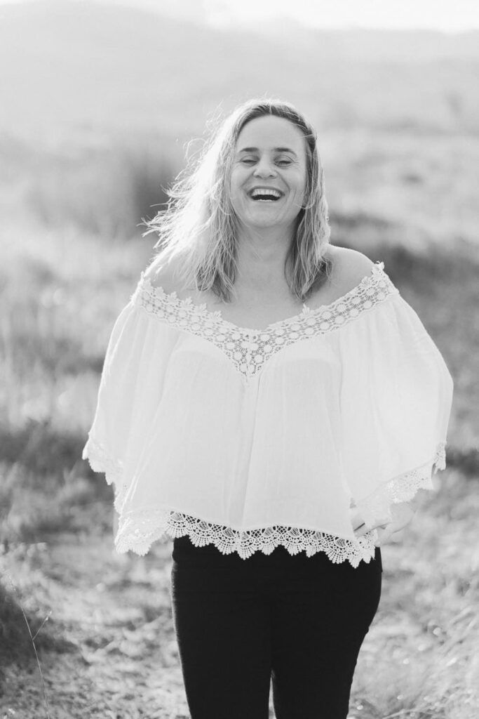 Black and white photo of a laughing woman in a white off-shoulder top standing in a field.