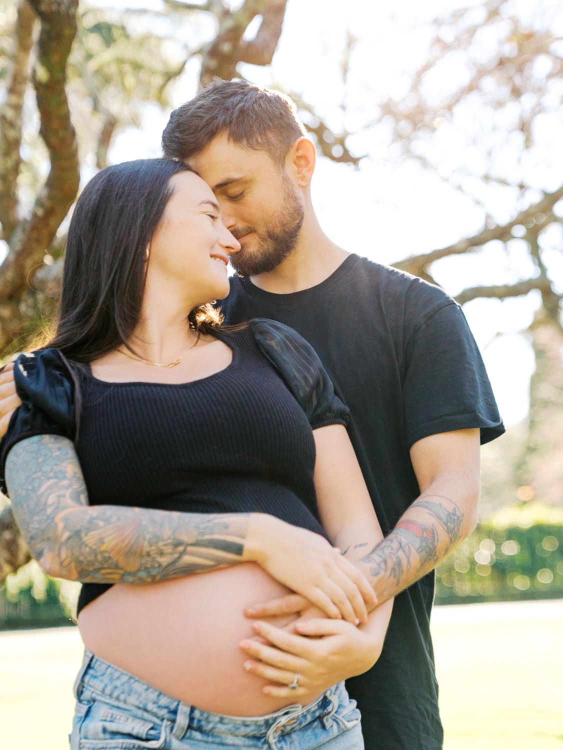 Pregnant couple embracing outdoors, both in black outfits with tattoos, sharing a tender moment under blooming trees.