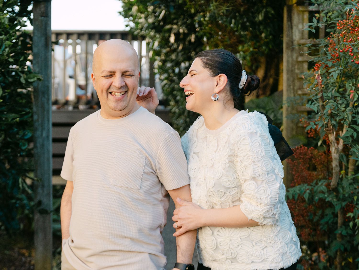 A laughing couple enjoying a sunny outdoor moment together, with lush greenery and a wooden fence in the background.