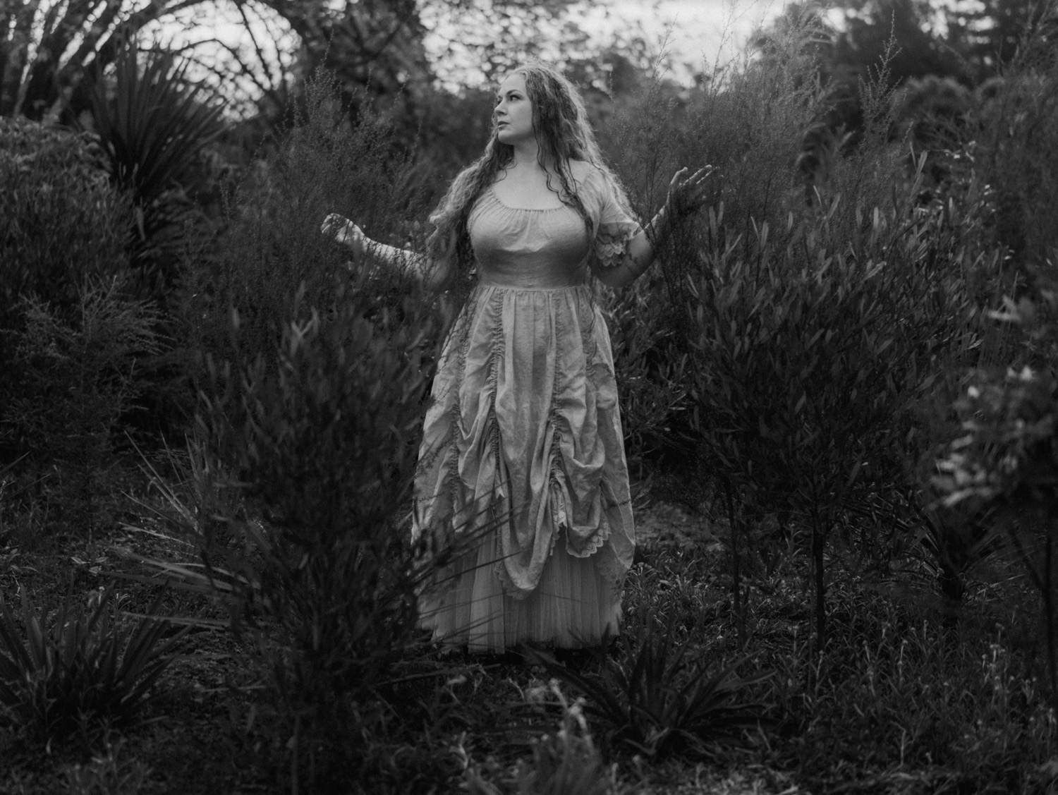 Black and white photo of a woman in a layered vintage dress among dense shrubs.