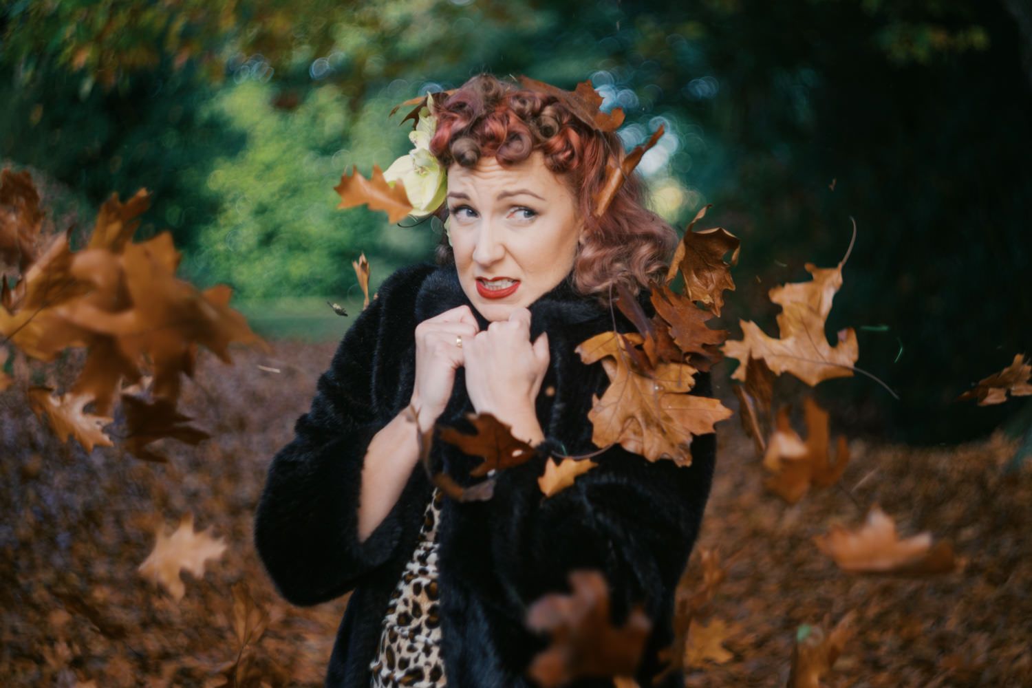 Woman with red hair poses among swirling autumn leaves in a forest, wearing a black coat and leopard print top.