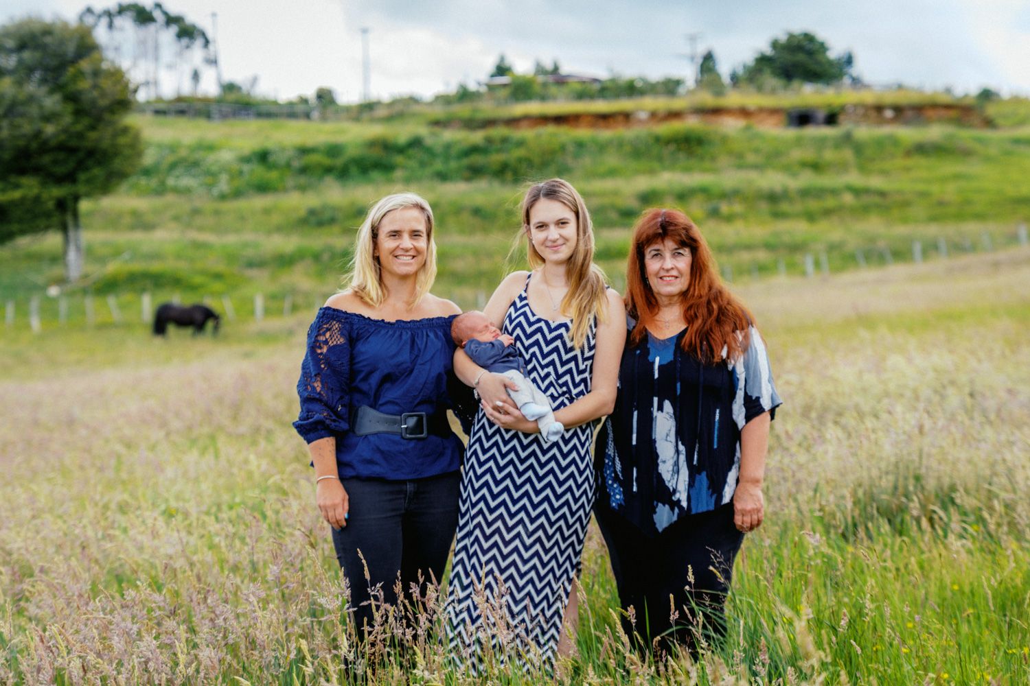 Three women smiling together in a lush green field with a fence and grazing animal in the background.