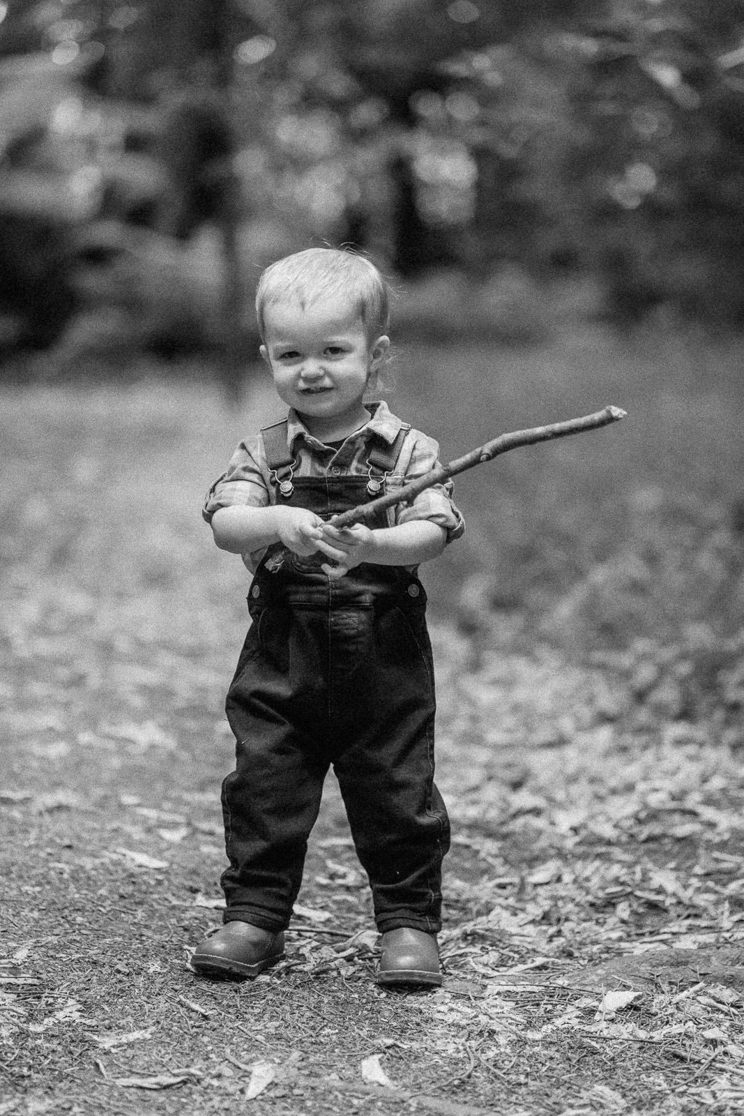 Black and white photo of a toddler boy in overalls holding a stick outdoors in a wooded park setting.