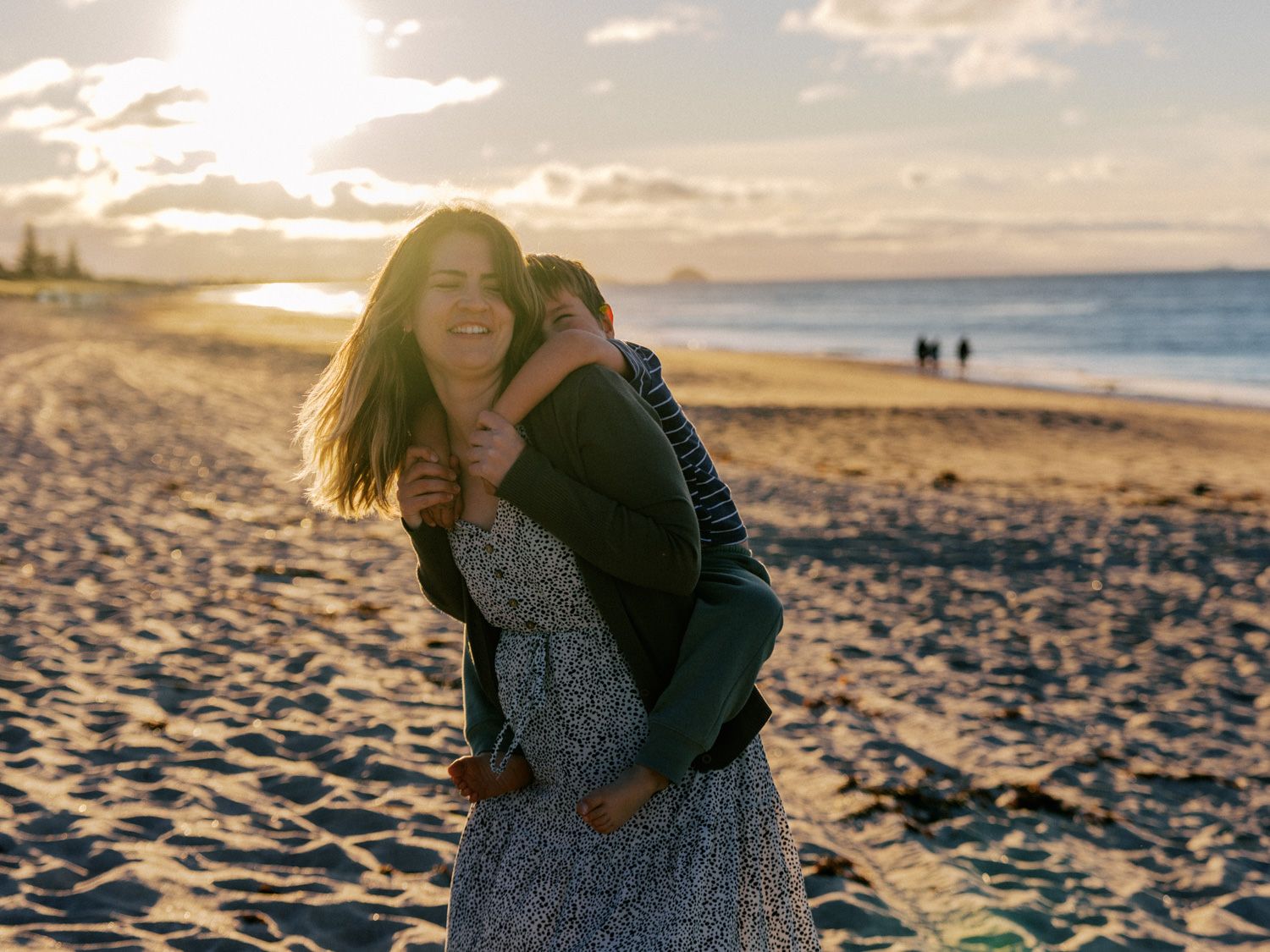 Couple embracing on a sandy beach at golden sunset, woman smiling joyfully, ocean and cloudy sky in background.