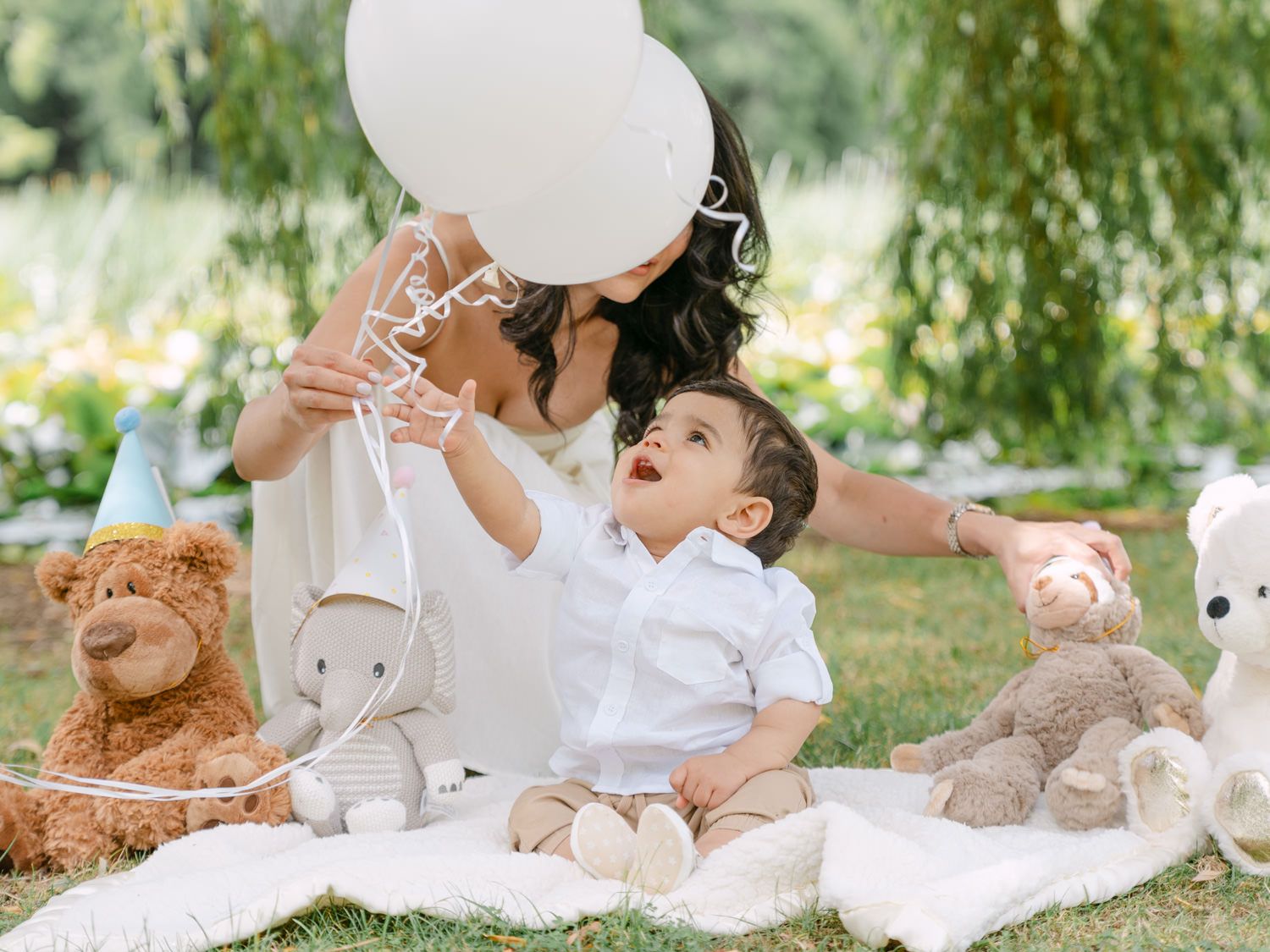 A mother and baby boy celebrate first birthday outdoors on a blanket with white balloons and stuffed animals.