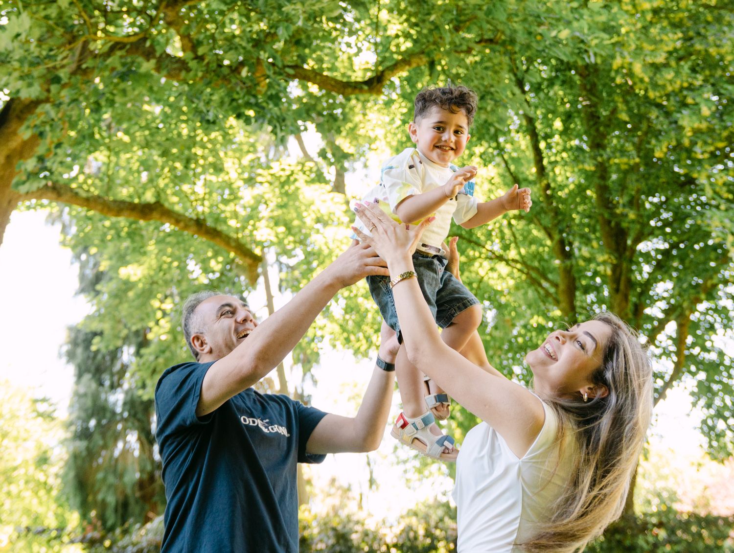 Happy family lifting a laughing toddler in the air outdoors, surrounded by lush green trees in a sunny park.