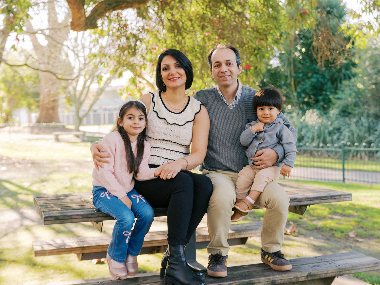 Family of four sitting on a picnic bench in a park, parents with two young children smiling outdoors.