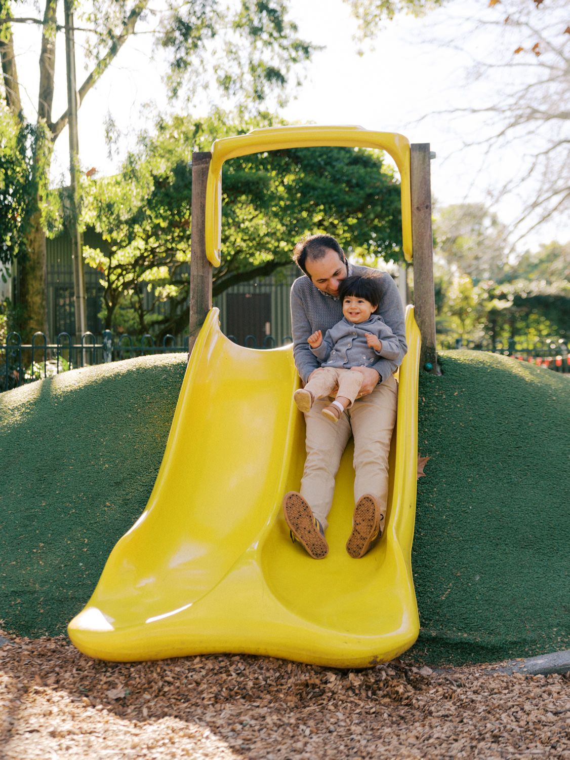 Father and child joyfully sliding down a yellow playground slide surrounded by lush green trees.