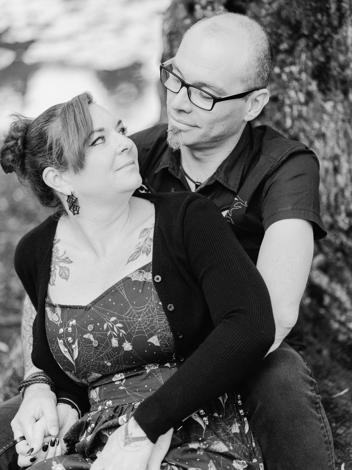 Black and white photo of a couple sitting together near a tree, woman with tattoos looking up at bespectacled man.