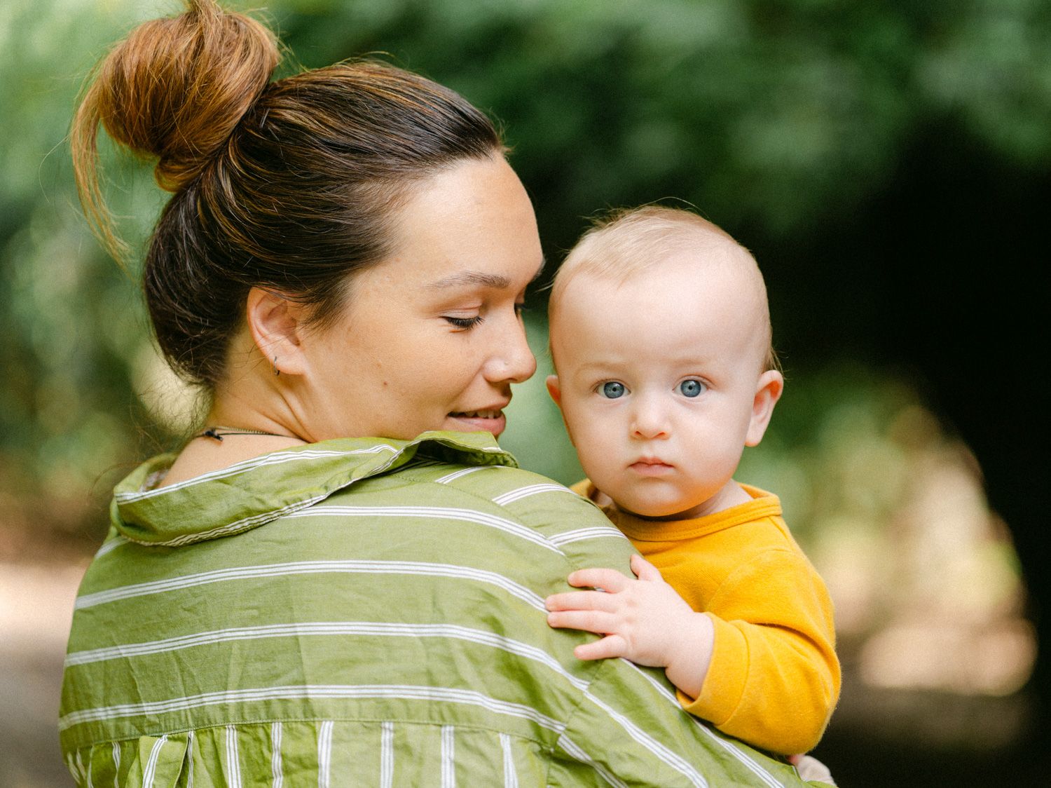 A mother in a green striped shirt holds her blue-eyed baby in a yellow onesie outdoors.