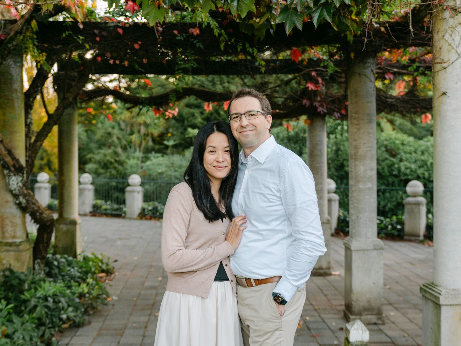 Couple posing under a floral pergola in a lush garden, woman in pink top and white skirt, man in light blue shirt.
