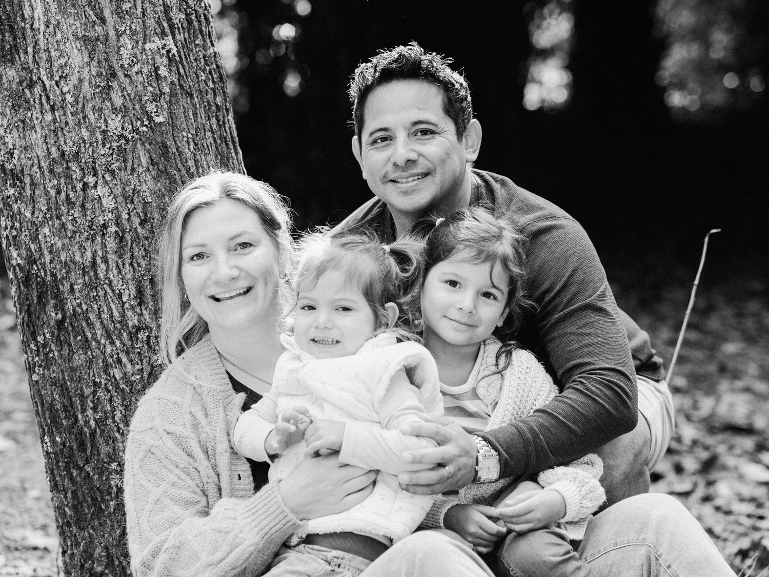 Black and white family portrait of parents with two young children sitting by a tree in a forest setting.