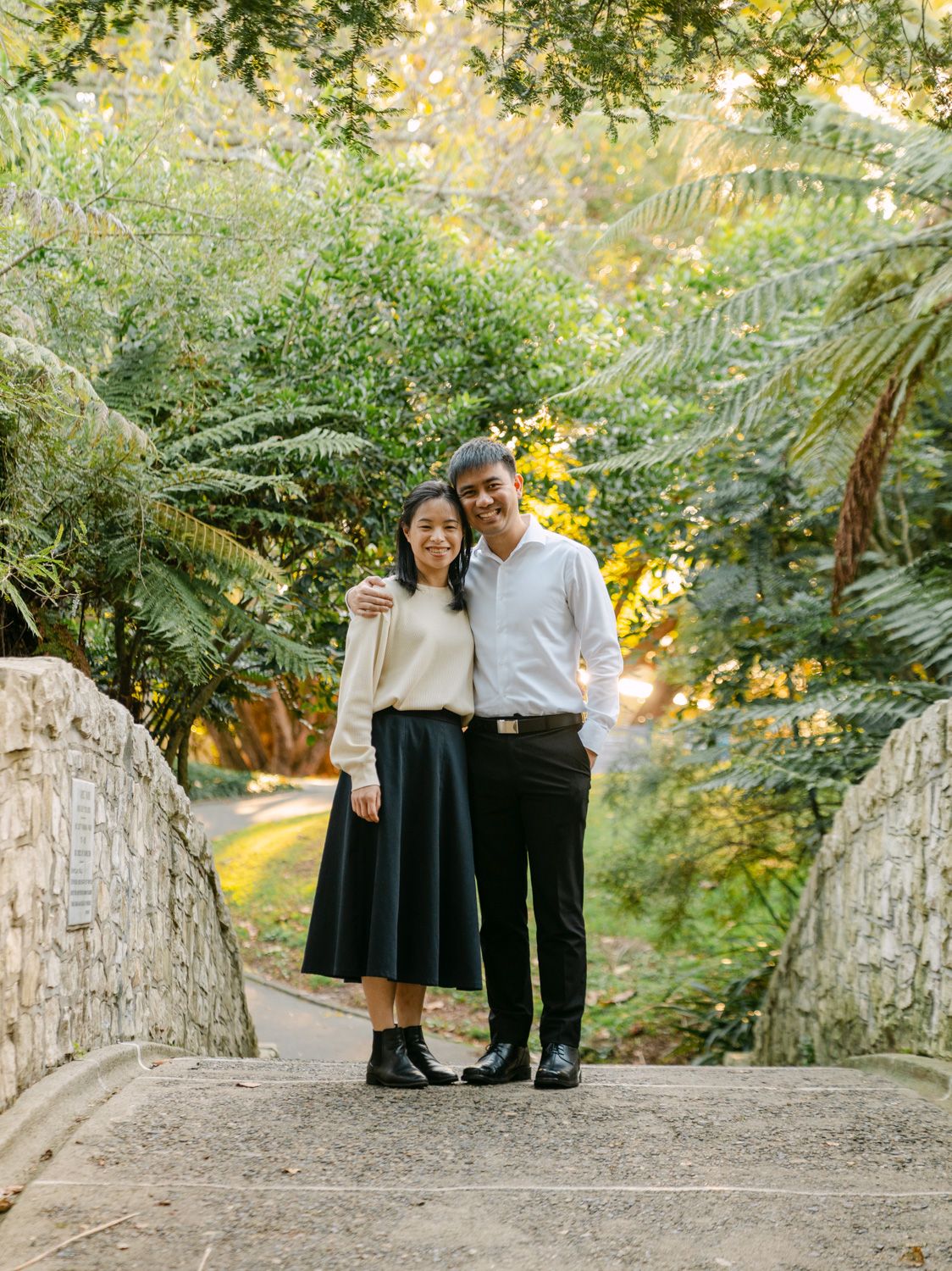Couple posing together on stone steps surrounded by lush green ferns and trees in a botanical garden.
