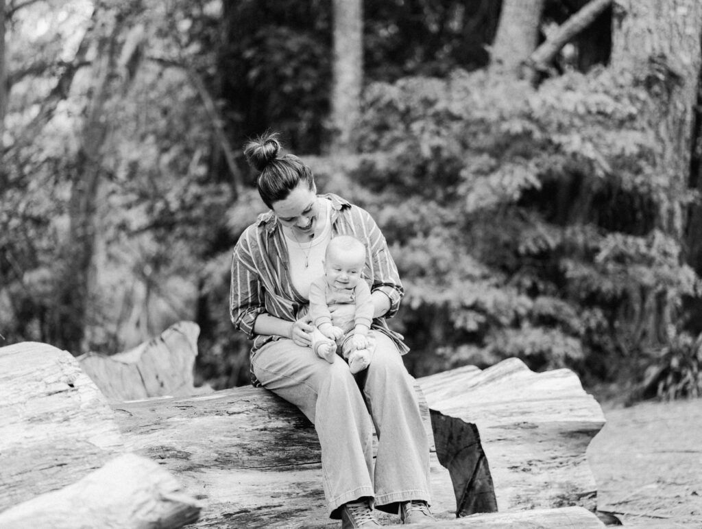 Black and white photo of a mother sitting on a log holding her baby in a forest setting.