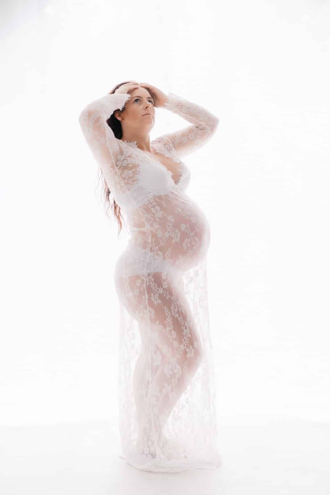 Pregnant woman in white lace gown posing elegantly against white background in maternity photo shoot.