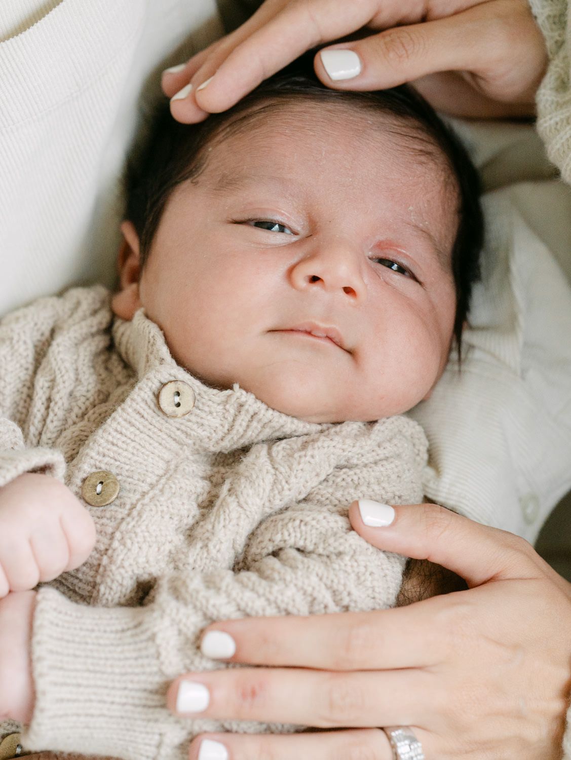 Newborn baby in beige knit cardigan cradled in mother's hands with white manicured nails.