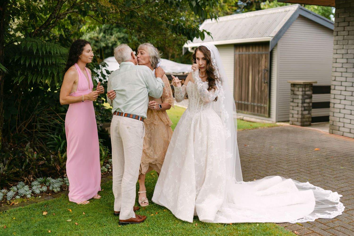 Bride in white lace gown with veil greets guests outdoors, woman in pink bridesmaid dress stands nearby.