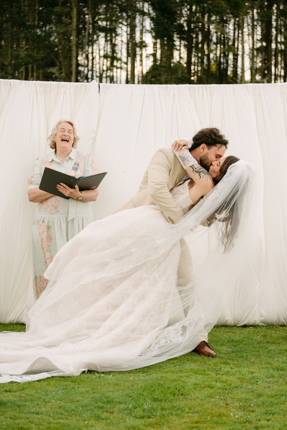 Groom dips bride for a kiss at outdoor wedding ceremony while officiant laughs behind white draped backdrop.