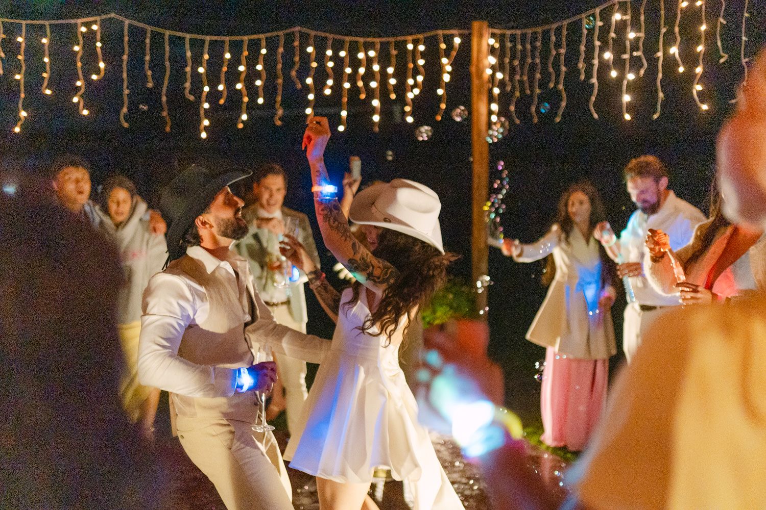 Guests dancing under string lights at an outdoor evening wedding reception with glowing accessories.