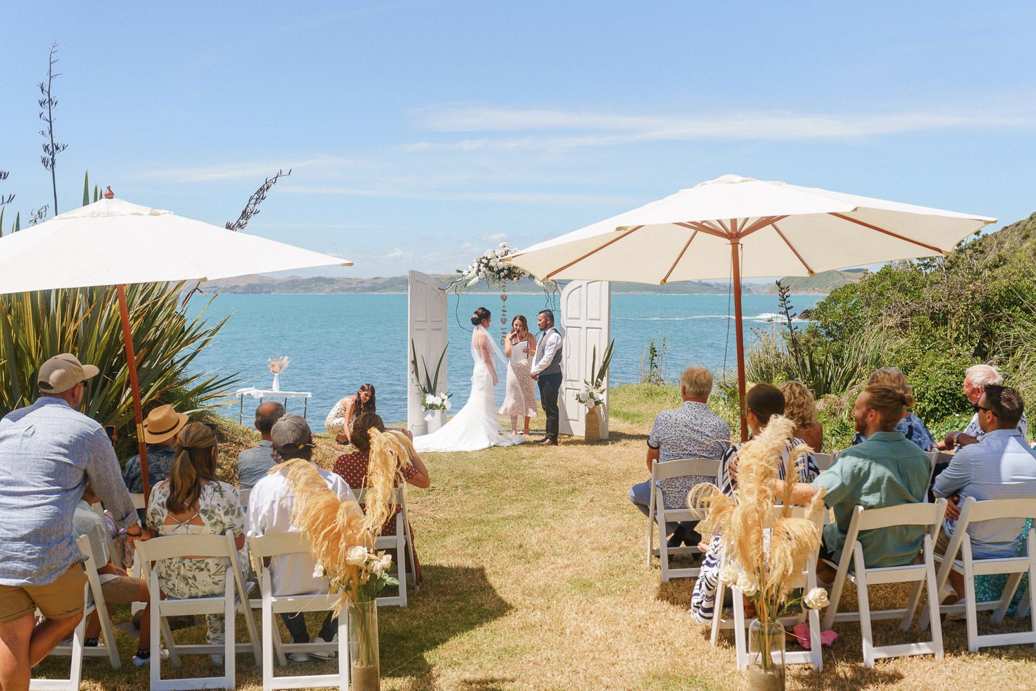 Outdoor beach wedding ceremony with guests seated on white chairs under umbrellas by the ocean on a sunny day.