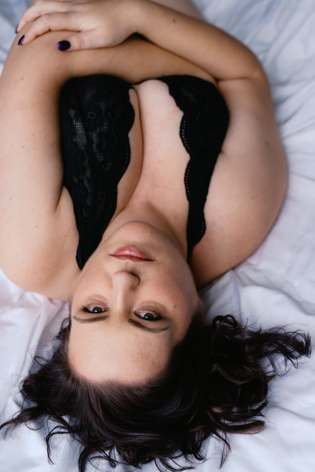Woman in black lace lingerie lying on white bedding, looking up at camera with dark curly hair spread out.