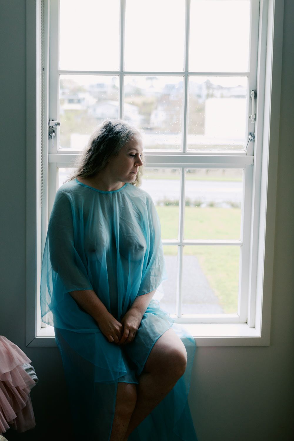 Woman in sheer blue robe sitting by a bright window in a serene boudoir photo.