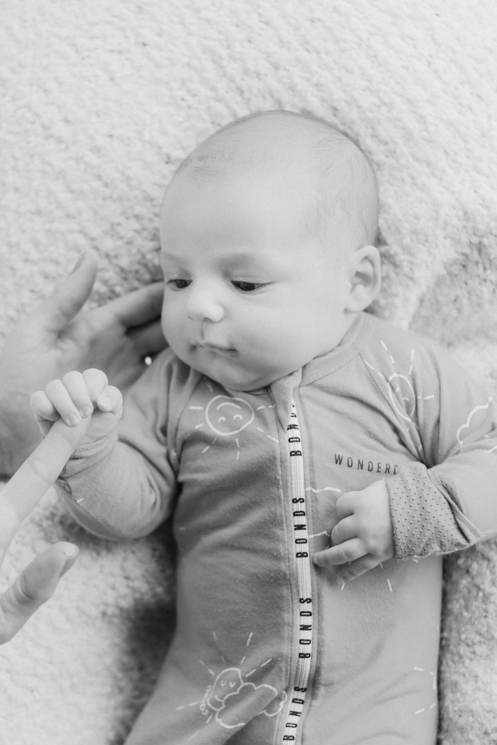 Black and white photo of a newborn baby in a Bonds zipsuit, gently holding an adult's finger while lying on a soft blanket.