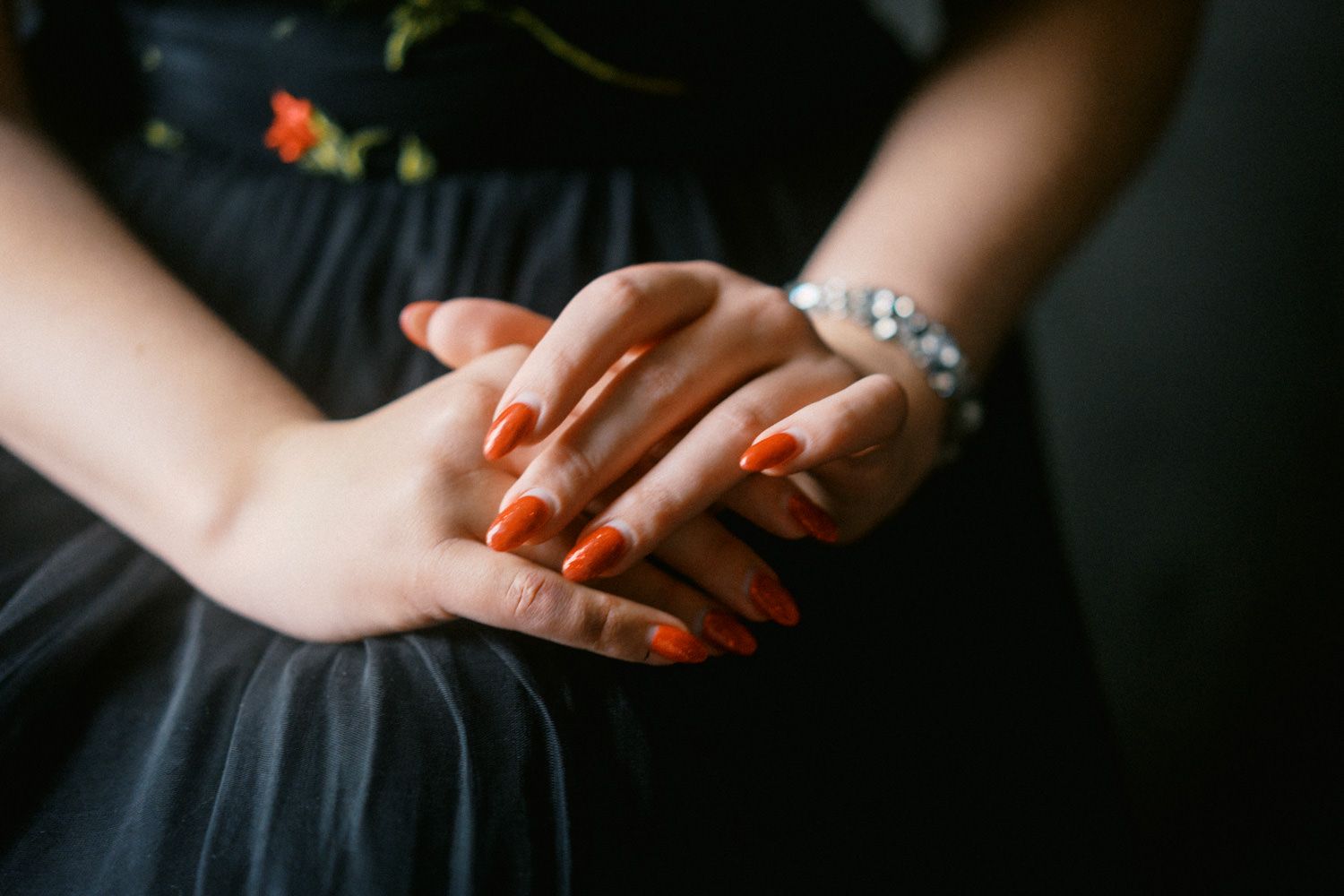 Close-up of woman's hands with orange-red nail polish and a crystal bracelet, resting on a black dress.