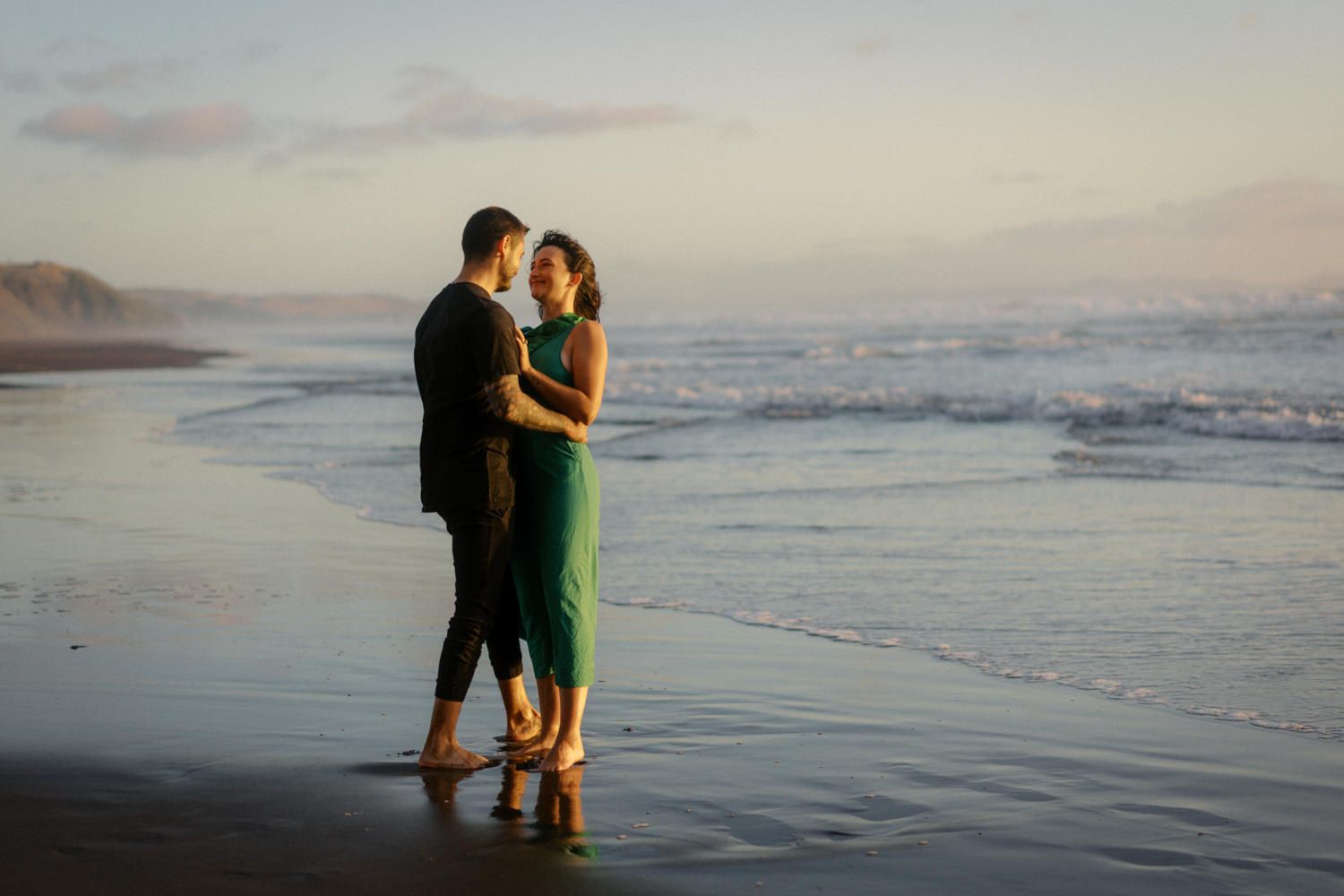 Couple embracing romantically on a sandy beach at sunset, waves crashing behind them, woman in green dress.