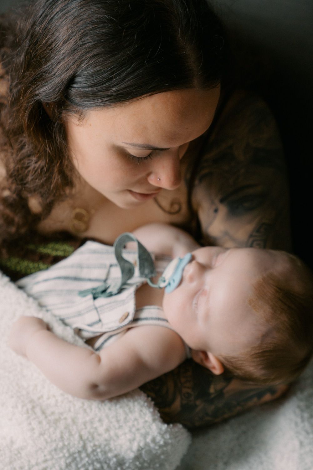 Tattooed mother gazing lovingly at her sleeping newborn baby dressed in a striped onesie with a pacifier.