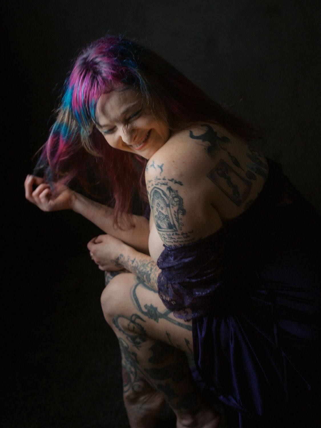 Tattooed woman with colorful pink and blue hair wearing a purple dress, smiling in a dark studio setting.