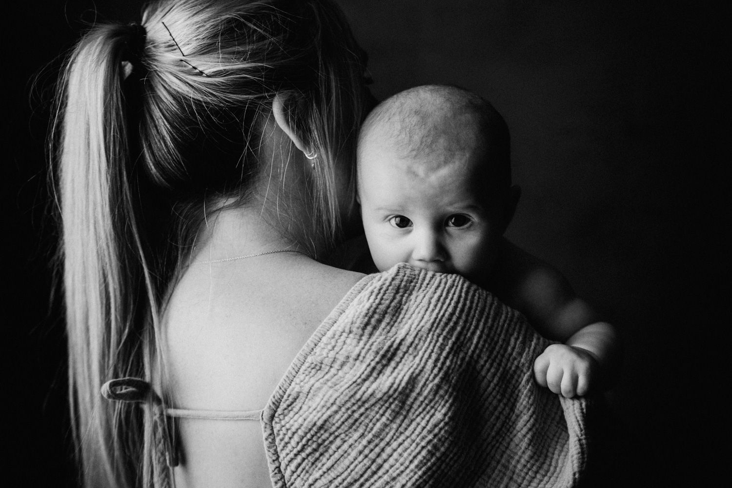 Black and white photo of a mother holding a newborn baby wrapped in a knit blanket over her shoulder.