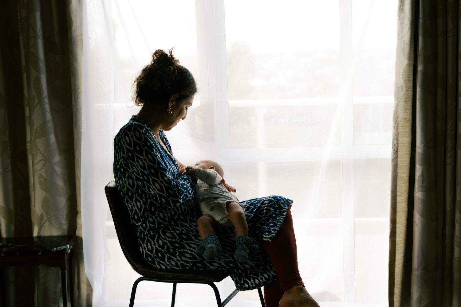 Silhouette of a mother sitting in a chair by a window, cradling her newborn baby in soft natural light.