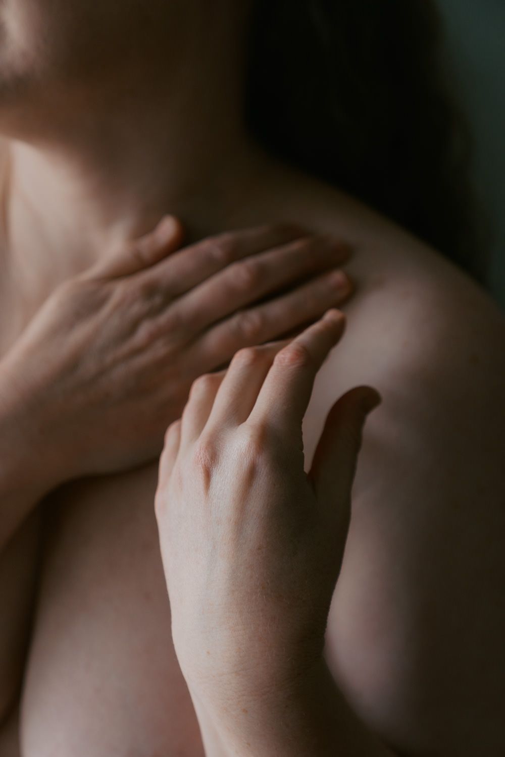 Close-up of hands gently resting on a person's chest and shoulder in soft, moody lighting.