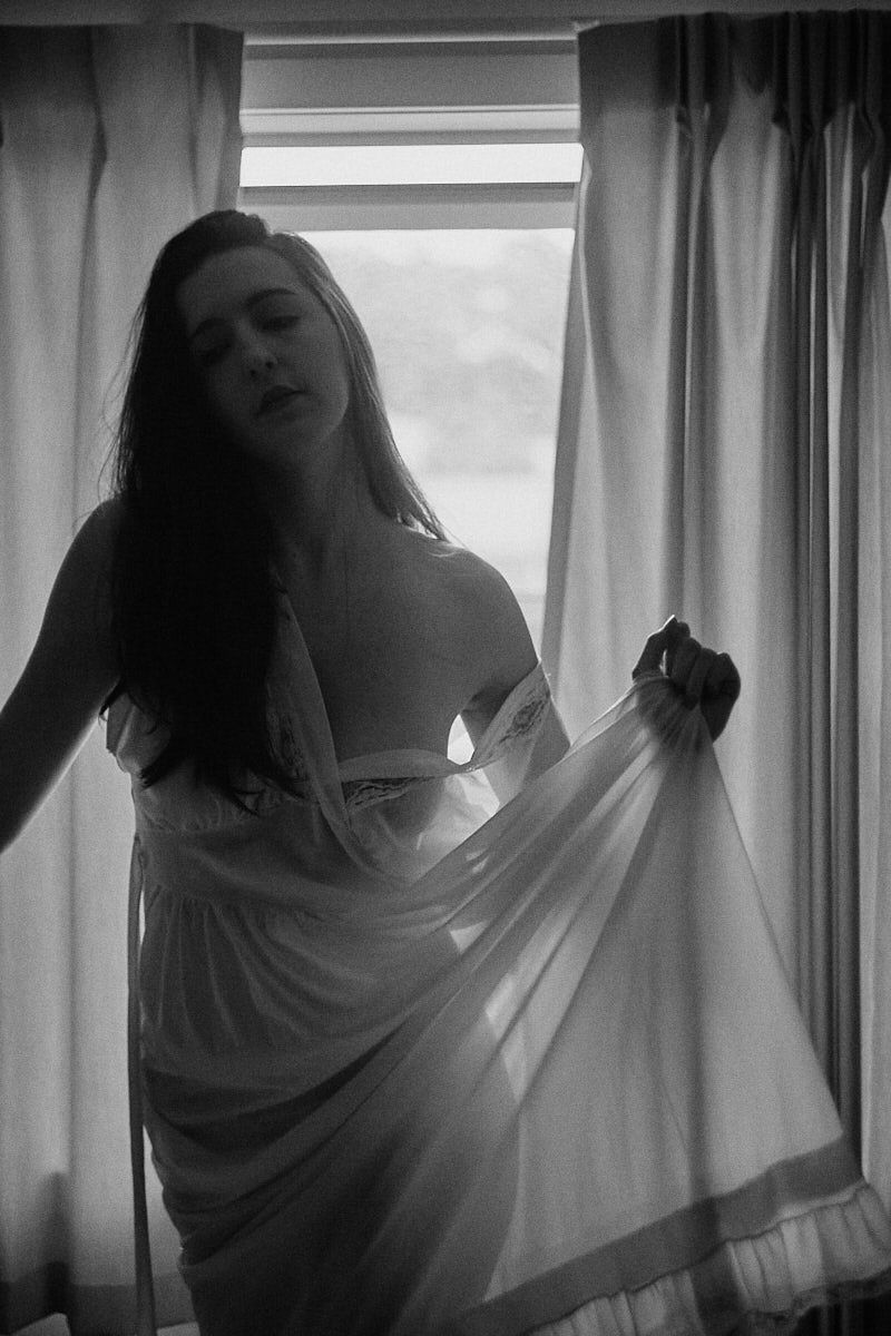 Black and white photo of a woman in sheer white lingerie posing by a sunlit window with curtains.