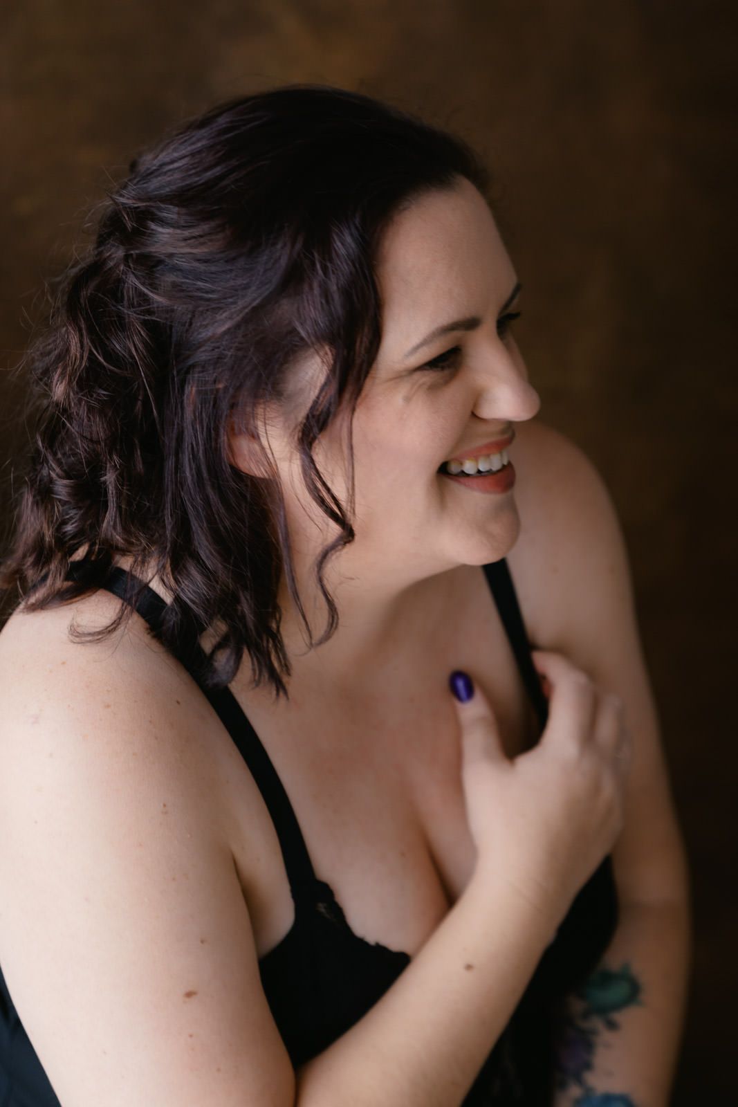 Smiling woman with dark hair wearing black lingerie, showcasing a tattoo on her arm in a studio setting.