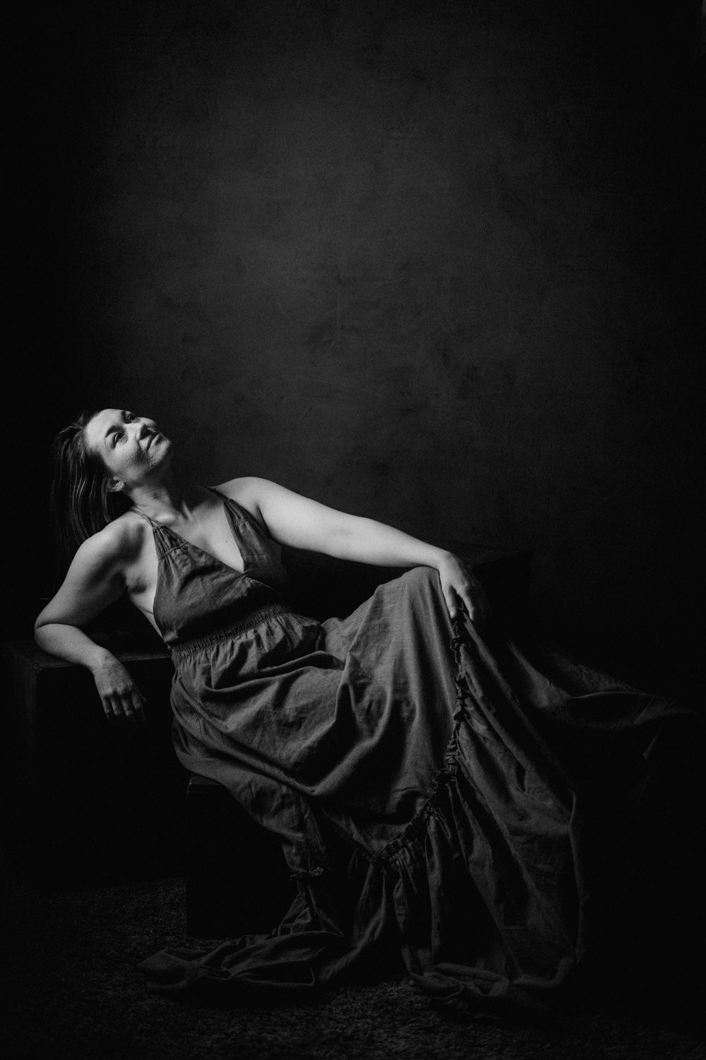 Black and white portrait of a woman in a flowing gown reclining gracefully on a chair against a dark backdrop.